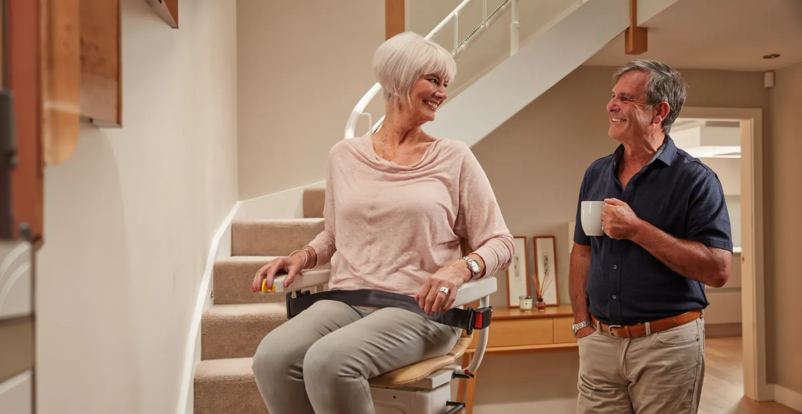 Frau sitzt auf einem Sitzlift an einer kurvigen Treppe, ihr Mann steht mit einer Tasse daneben